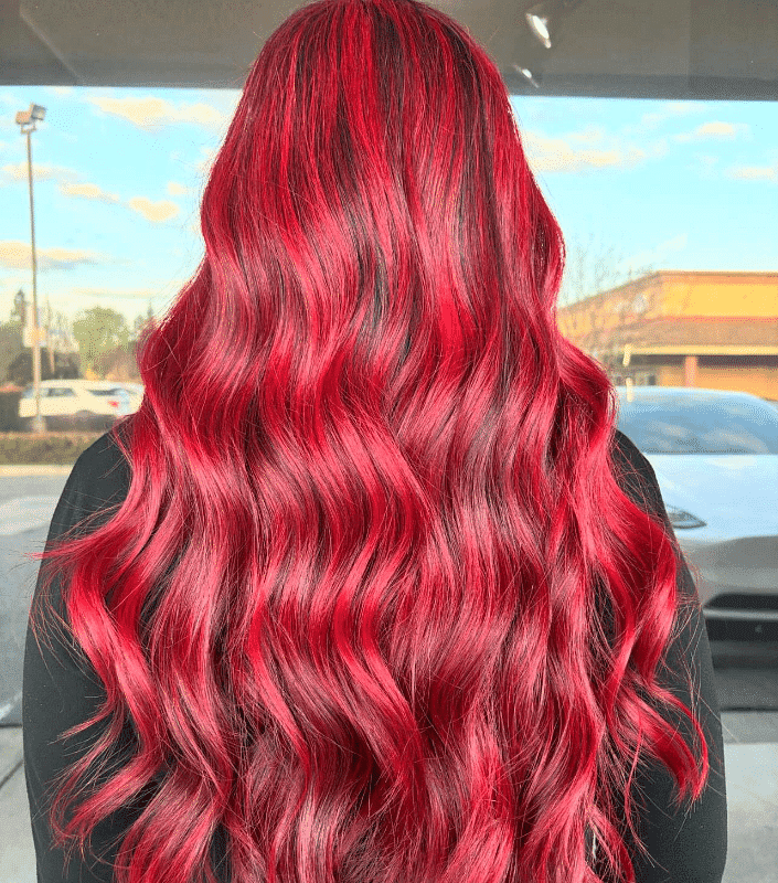 Woman with long, wavy, vibrant red hair standing outdoors with cars in the background.