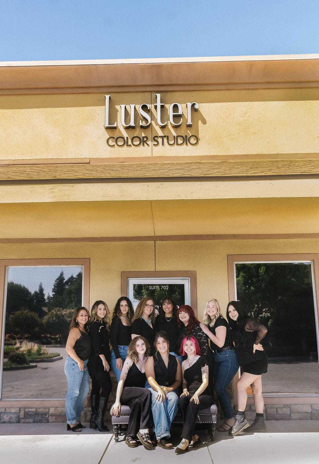 Group of twelve people posing in front of Luster Color Studio on a sunny day.